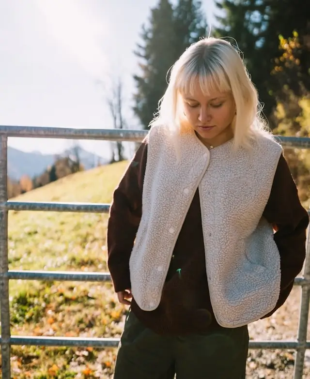 Young woman with blonde hair wearing a white fleece vest and dark long sleeves, standing outdoors near a metal fence with trees and hills in the background.