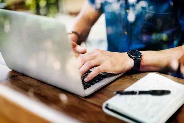 Person wearing a watch typing on a laptop at a wooden desk with a notebook and pen nearby.