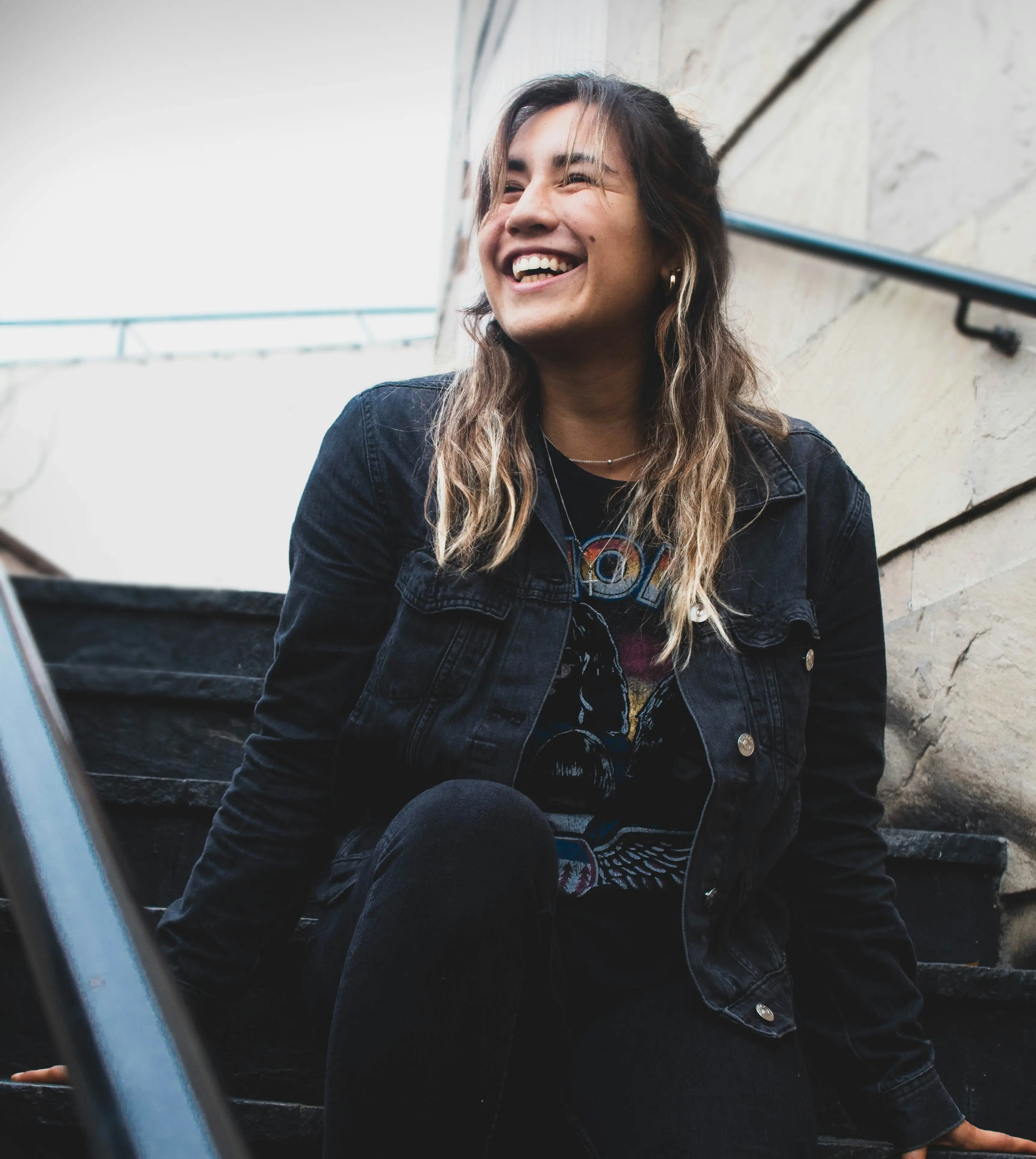 Young woman with long hair smiling while sitting on outdoor stone steps wearing a black denim jacket and graphic t-shirt.