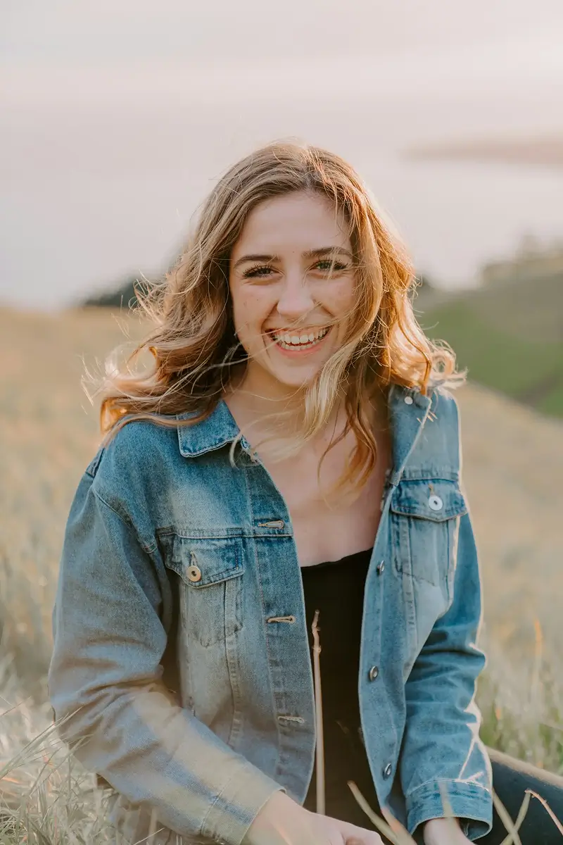 Young woman with long wavy hair smiling outdoors in a field wearing a denim jacket.