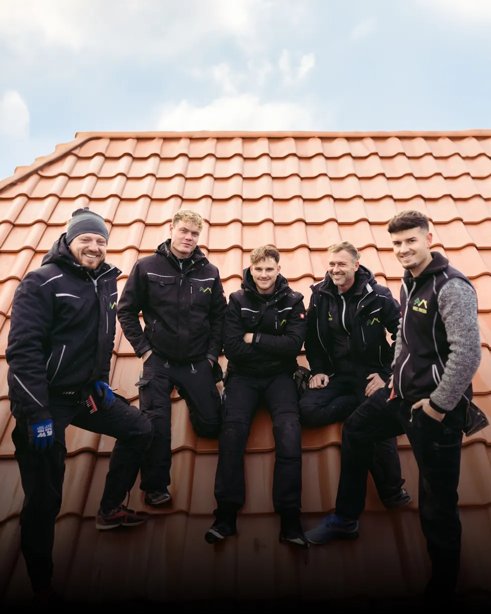 Fünf Männer in schwarzer Arbeitskleidung sitzen und stehen auf einem Ziegeldach und lächeln in die Kamera.