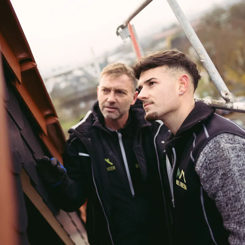 Zwei Handwerker inspizieren ein Dach, einer zeigt auf Dachziegel.