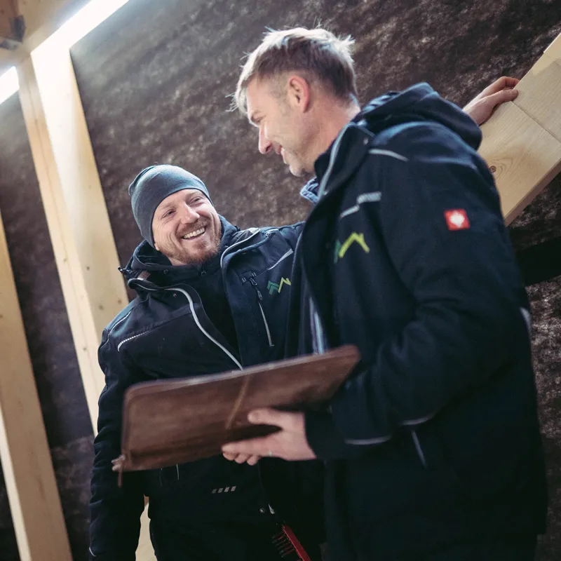 Zwei Männer in Arbeitsjacken unterhalten sich lachend auf einer Baustelle, einer hält ein Leder-Tablet-Etui.