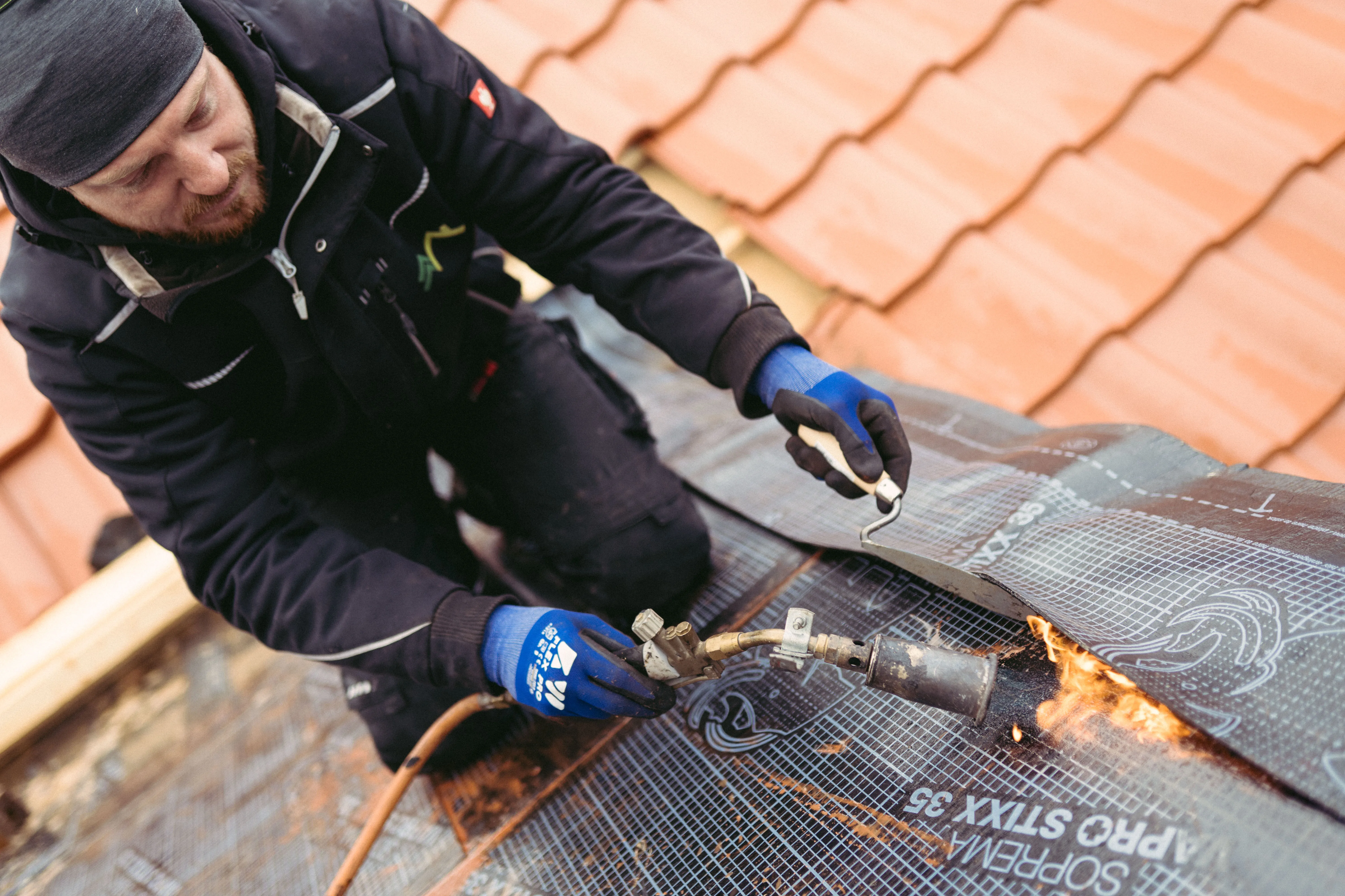 Handwerker mit blauen Handschuhen bringt Dachbahnen mit Brenner auf einem Dach mit roten Dachziegeln an.