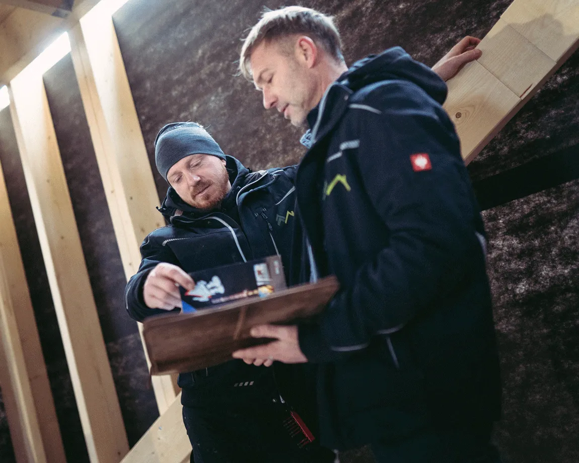 Zwei Männer in Arbeitskleidung prüfen Baupläne in einem Holzrahmenbau.