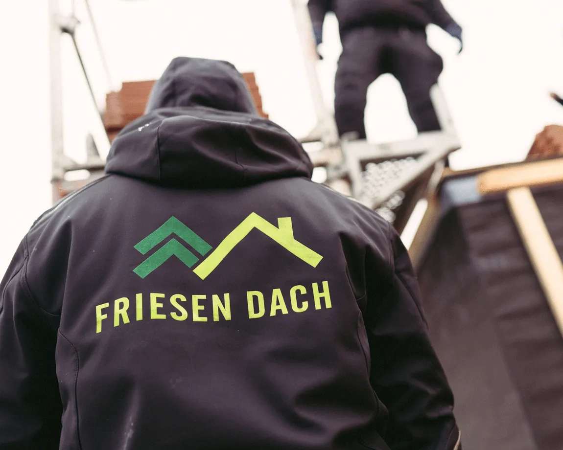 Person trägt eine schwarze Jacke mit dem Logo und Schriftzug 'Friesen Dach' auf dem Rücken, vor einer Baustelle mit Gerüst und einem weiteren Arbeiter.