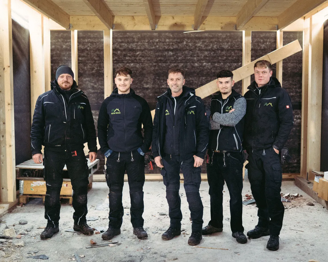 Fünf Handwerker in schwarzer Arbeitskleidung stehen in einem unvollendeten Holzrahmenbau.