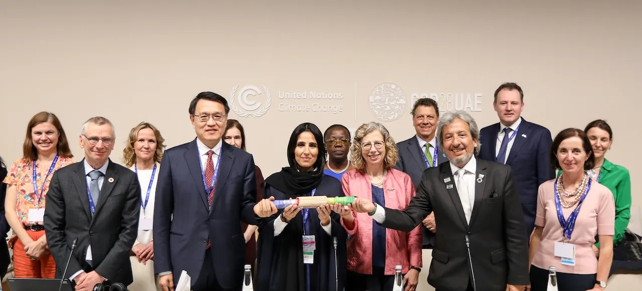 A group of around fourteen international delegates and leaders, including men and women in formal attire, stand in a line facing the camera. The backdrop features the United Nations Climate Change and COP28 UAE logos. In the center, a woman in a black abaya and headscarf and a man in a dark suit hold a long, segmented, cylindrical object between them. The group is smiling. This image captures a moment related to the COP28 Joint Statement on Climate, Nature and People.