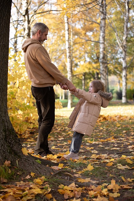 Man holding hands with young girl leaning back playfully in an autumn park with yellow leaves on the ground.