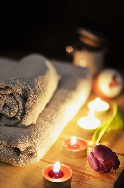 Rolled and folded beige towels beside lit candles and a purple tulip on a wooden surface.