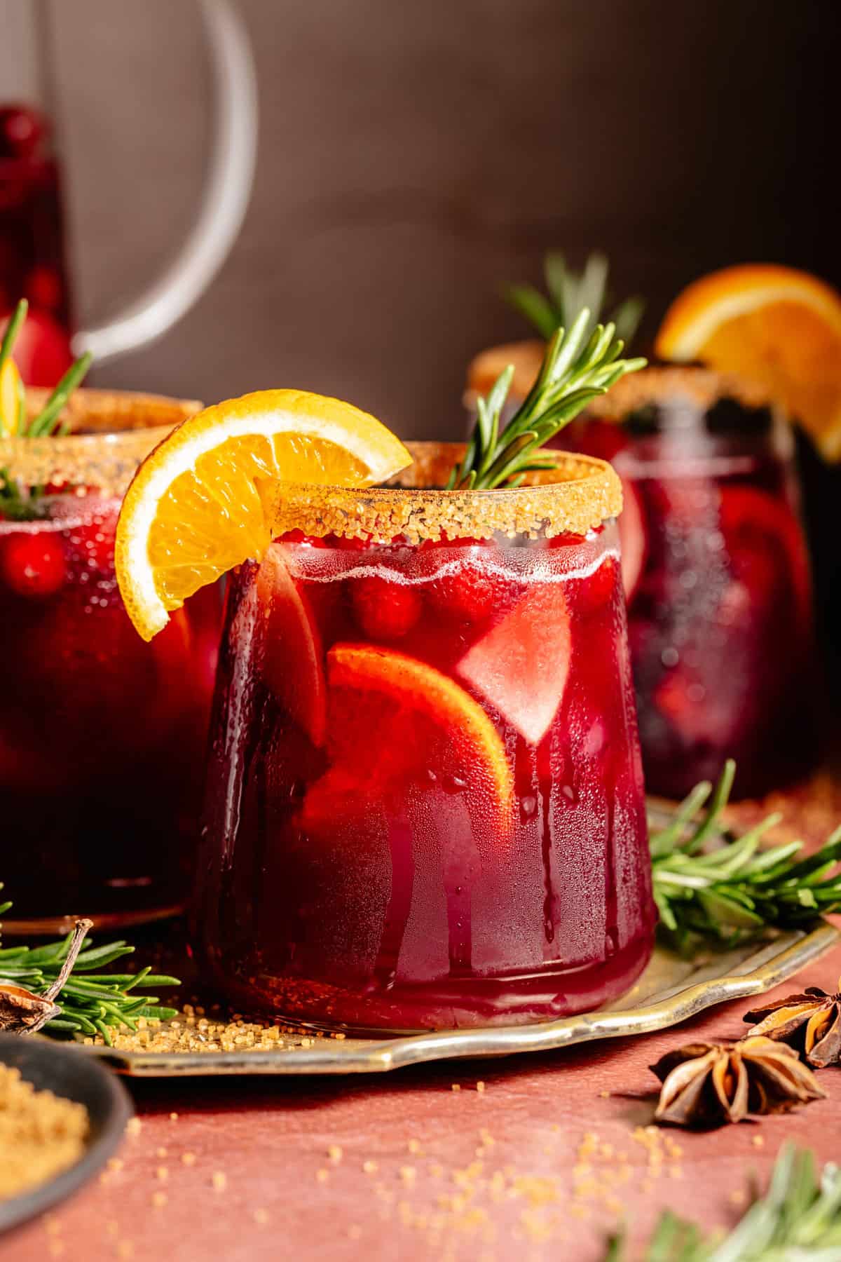 Glass of red sangria garnished with orange slice and rosemary sprig on a tray.
