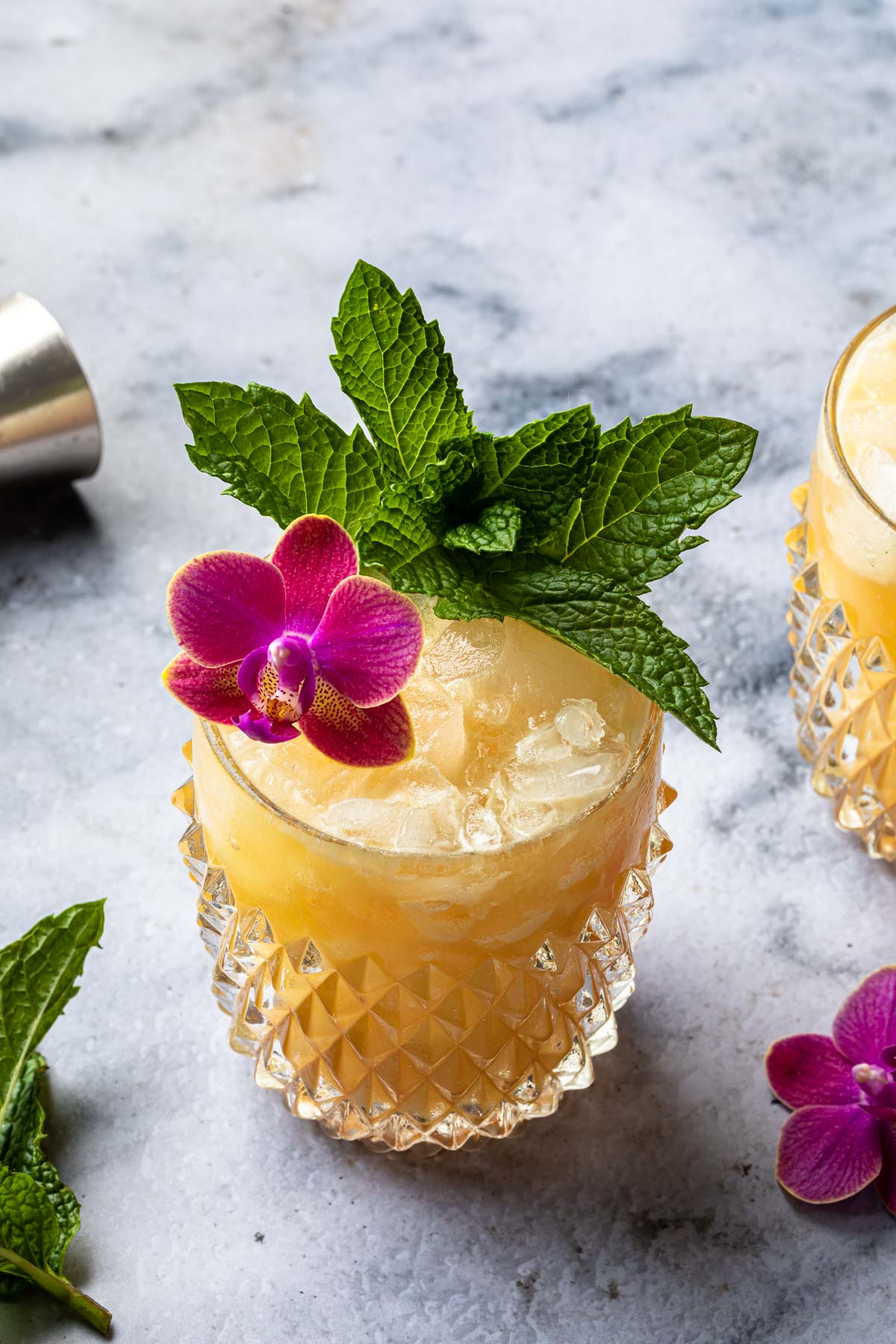 Tropical iced cocktail garnished with fresh mint leaves and a purple orchid flower in a textured glass on a marble surface.