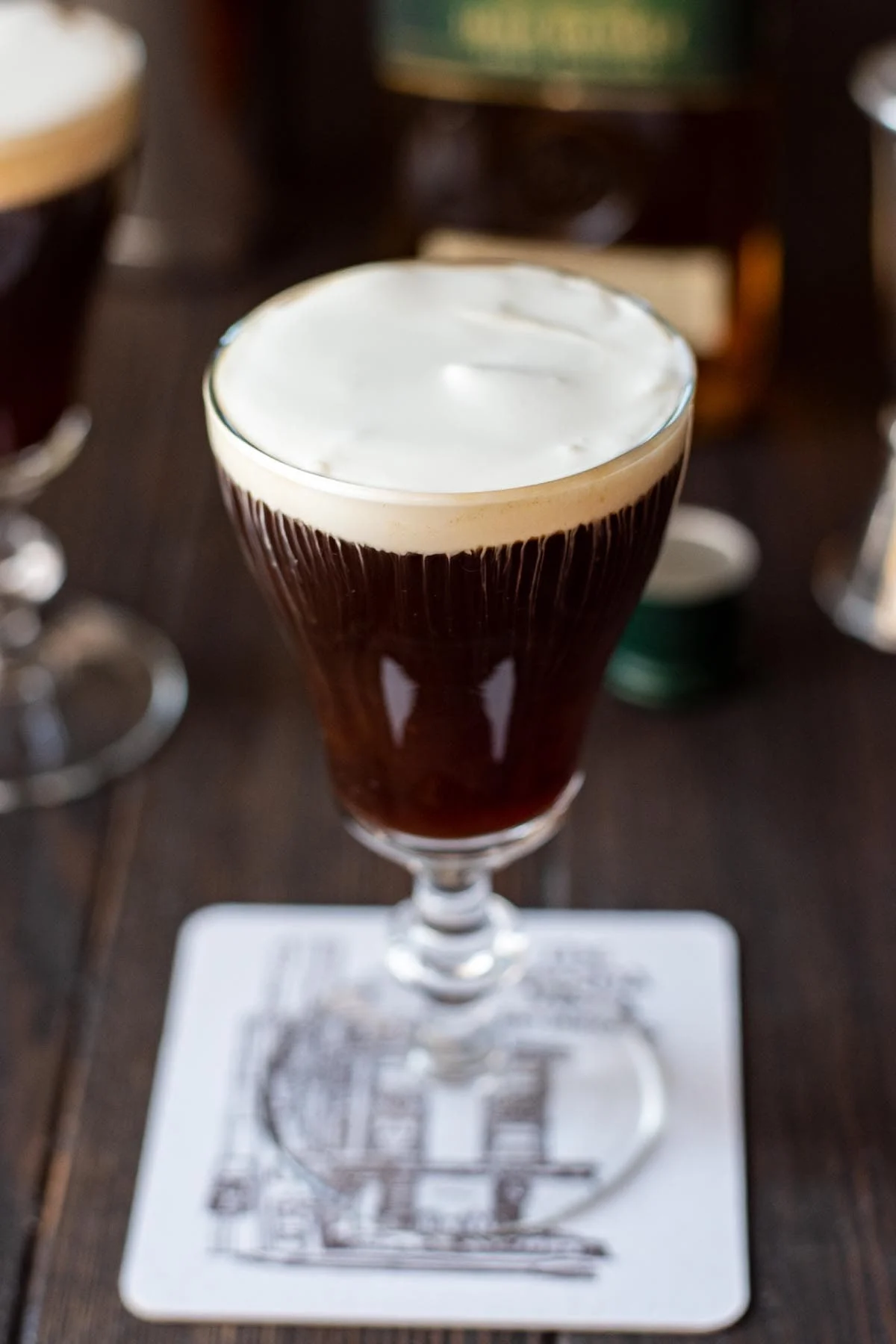 Close-up of a glass of Irish coffee topped with thick cream, placed on a coaster on a wooden table.