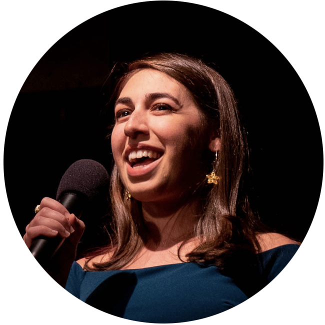 Young woman with brown hair and gold flower earrings smiling and speaking into a microphone against a dark background.