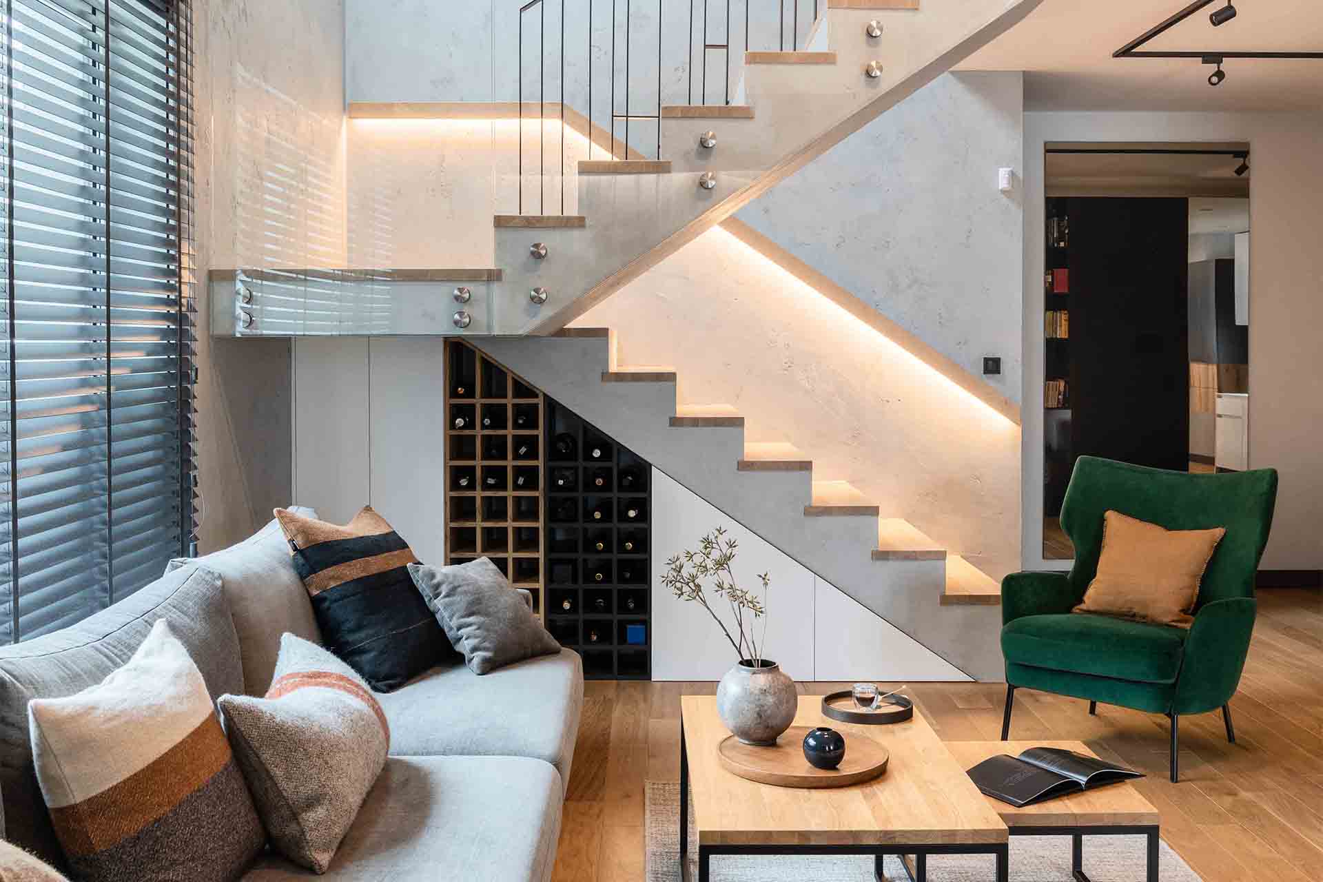 Modern living room with gray sofa, green armchair, wooden coffee tables, and stairs with built-in wine rack and LED lighting.