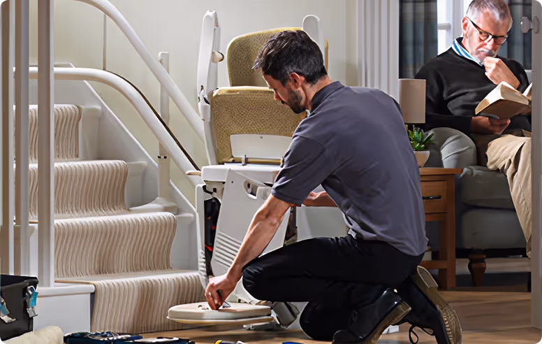Technician repairing a residential stairlift while the homeowner sits nearby.