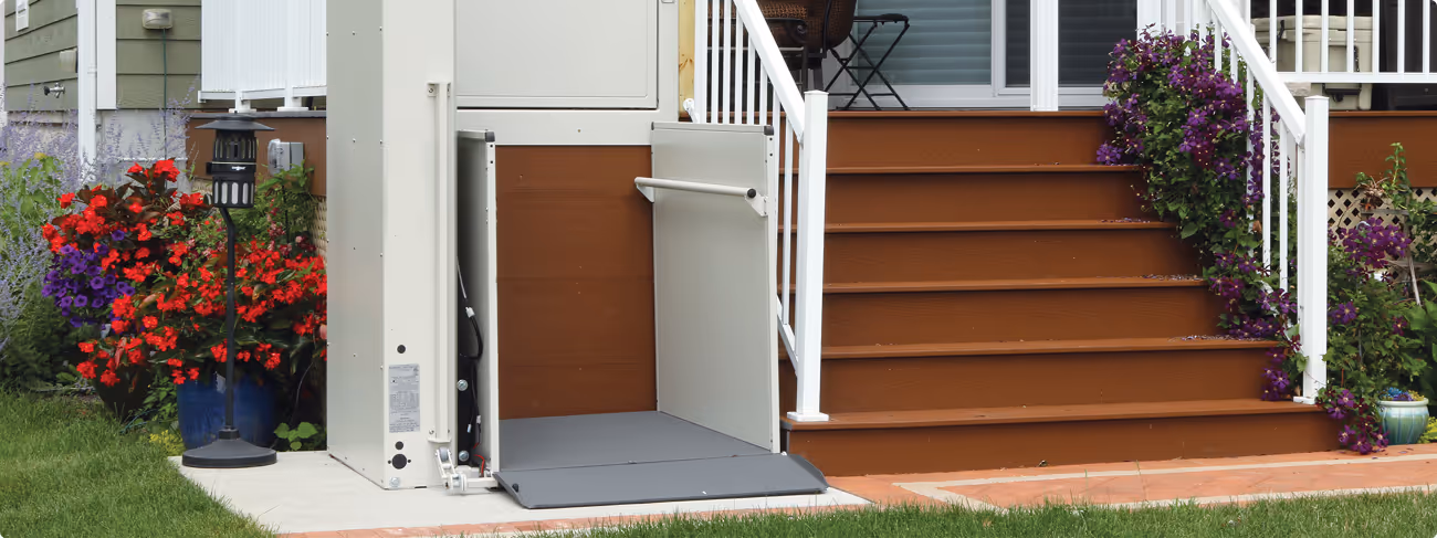 Outdoor wheelchair lift next to wooden brown stairs with white railing and flowering plants.