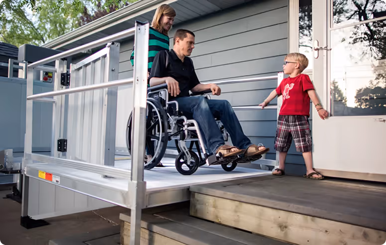 Man in a wheelchair on a wheelchair lift ramp outside a house with a woman standing behind him and a young boy nearby.