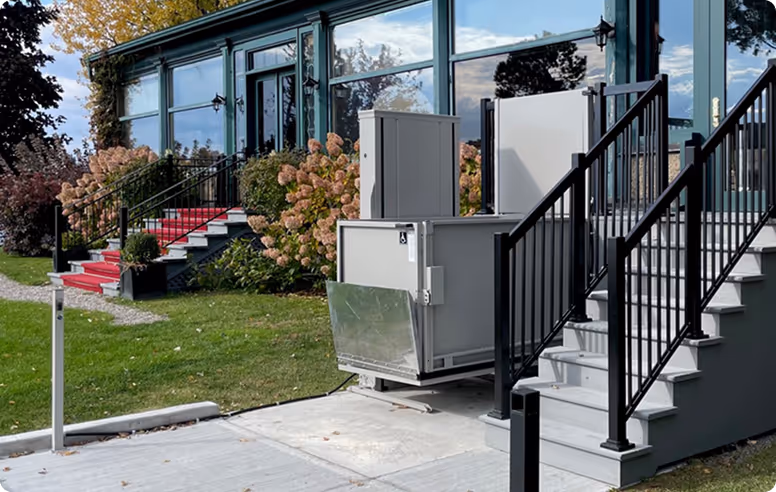 Outdoor residential wheelchair lift installed next to stairs with black railing and a glass building facade in the background.