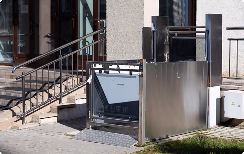 Outdoor wheelchair lift installed next to stairs of a building entrance, with metal railings and a textured ramp platform.