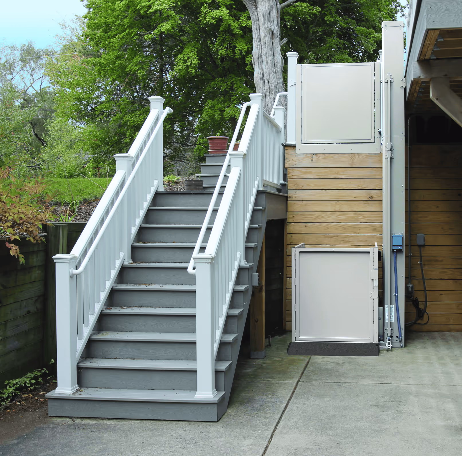 Outdoor wooden staircase with white railings next to a residential vertical wheelchair lift on concrete ground.