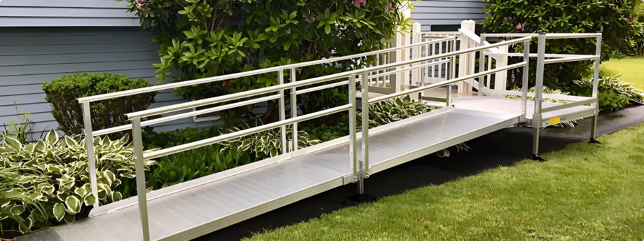 Metal wheelchair ramp with handrails leading to a house entrance surrounded by greenery and lawn.