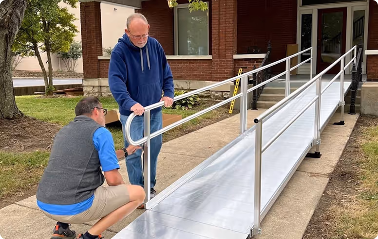 Two men assembling a metal wheelchair ramp outside a brick building.