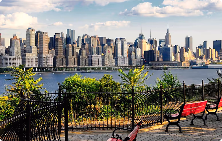 View of a city skyline across a river from a park with red benches and black wrought iron fencing.