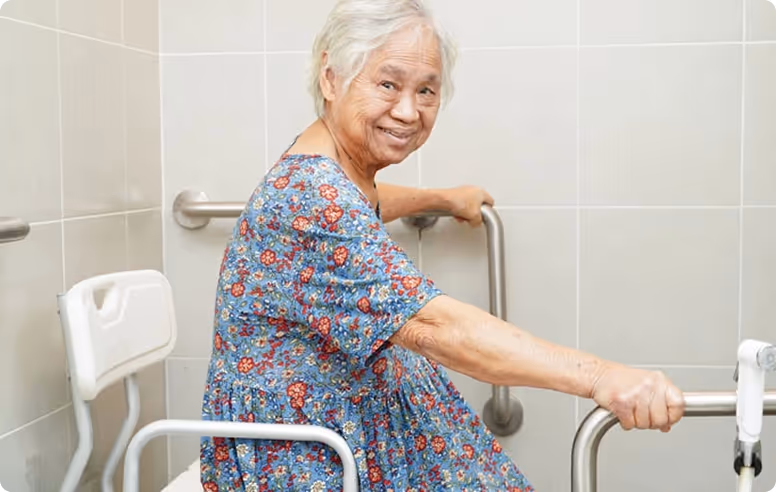Smiling elderly woman sitting on a shower chair holding onto bathroom grab bars.