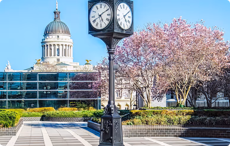Black antique street clock showing 10:10 in a plaza with pink blossoming trees and a domed building in the background under a clear blue sky.