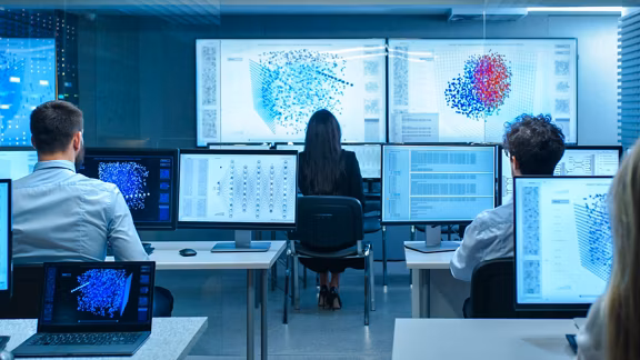 Team of data scientists analyzing 3D data visualizations and code on multiple computer monitors in a control room environment.