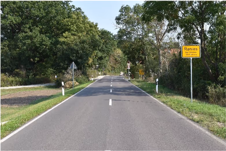 Straight rural road with white dashed center lines flanked by green trees and a yellow town sign reading 'Ranies' on the right.