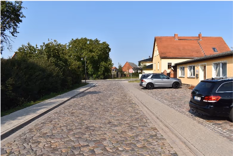 Cobblestone street lined with trees on the left and parked cars beside houses with red roofs on the right under a clear blue sky.