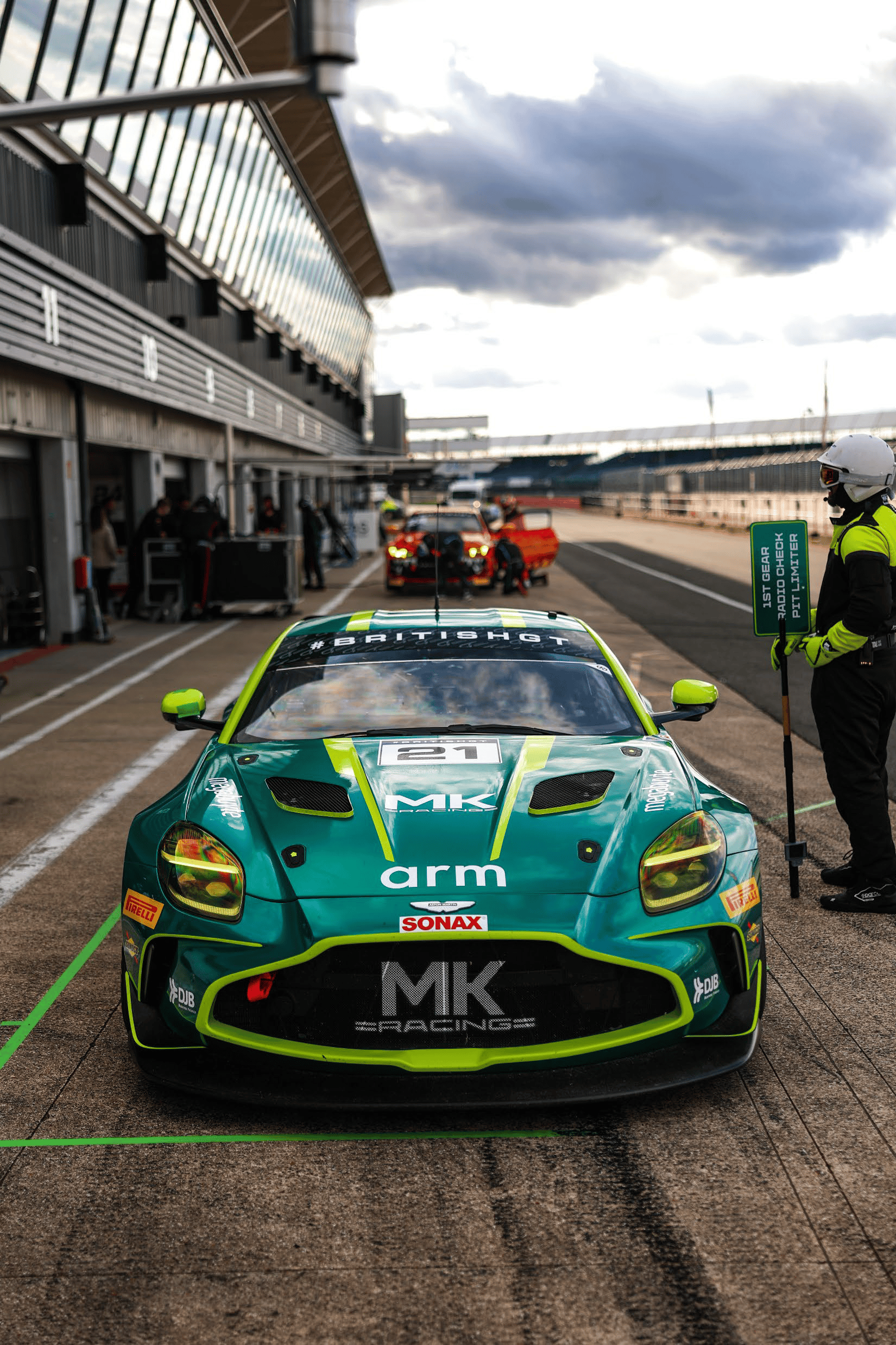 Green and yellow race car #21 in pit lane with team member holding 1st gear sign beside it.