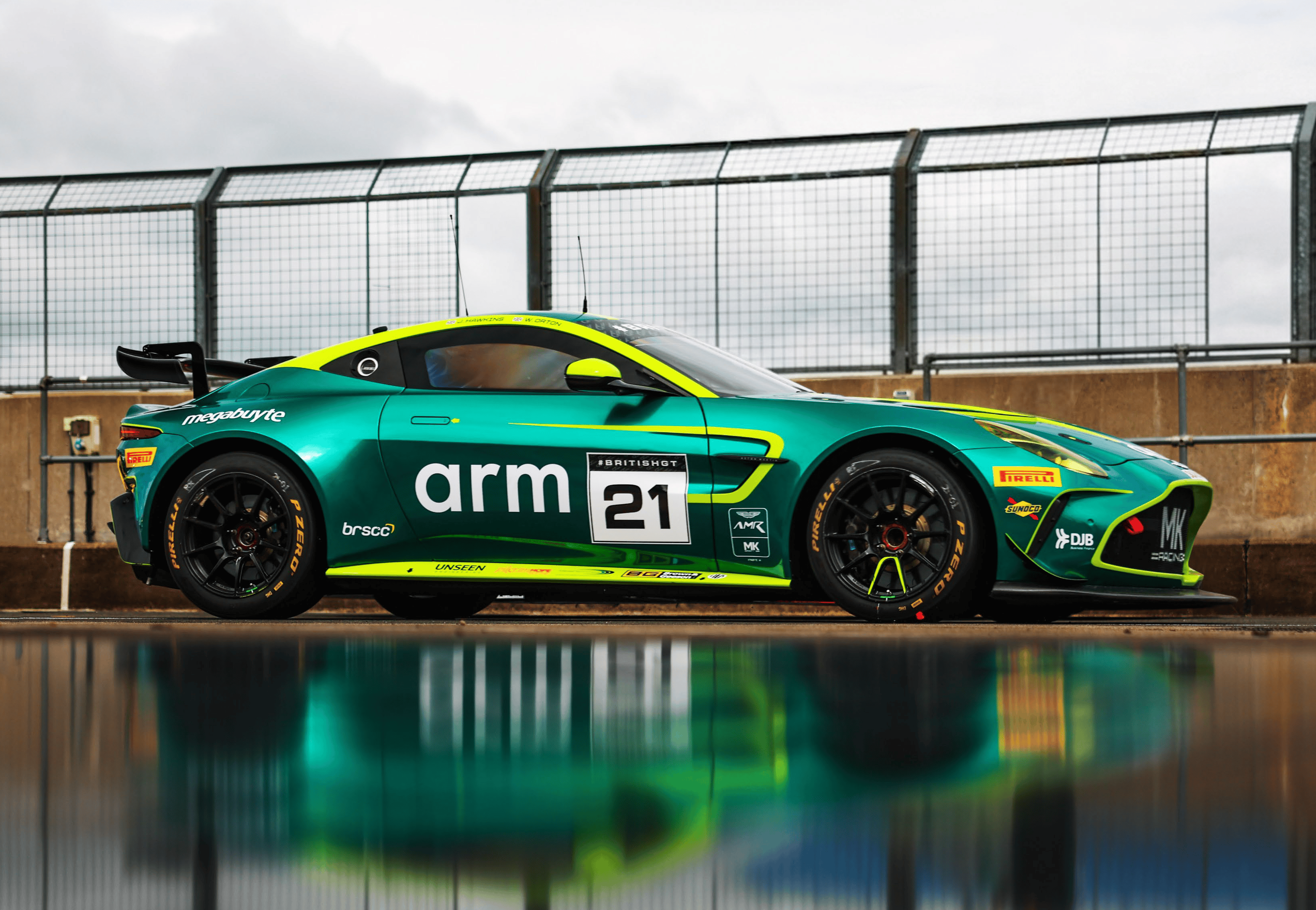 Green and yellow race car with number 21 and arm logo, parked in front of a wire fence.