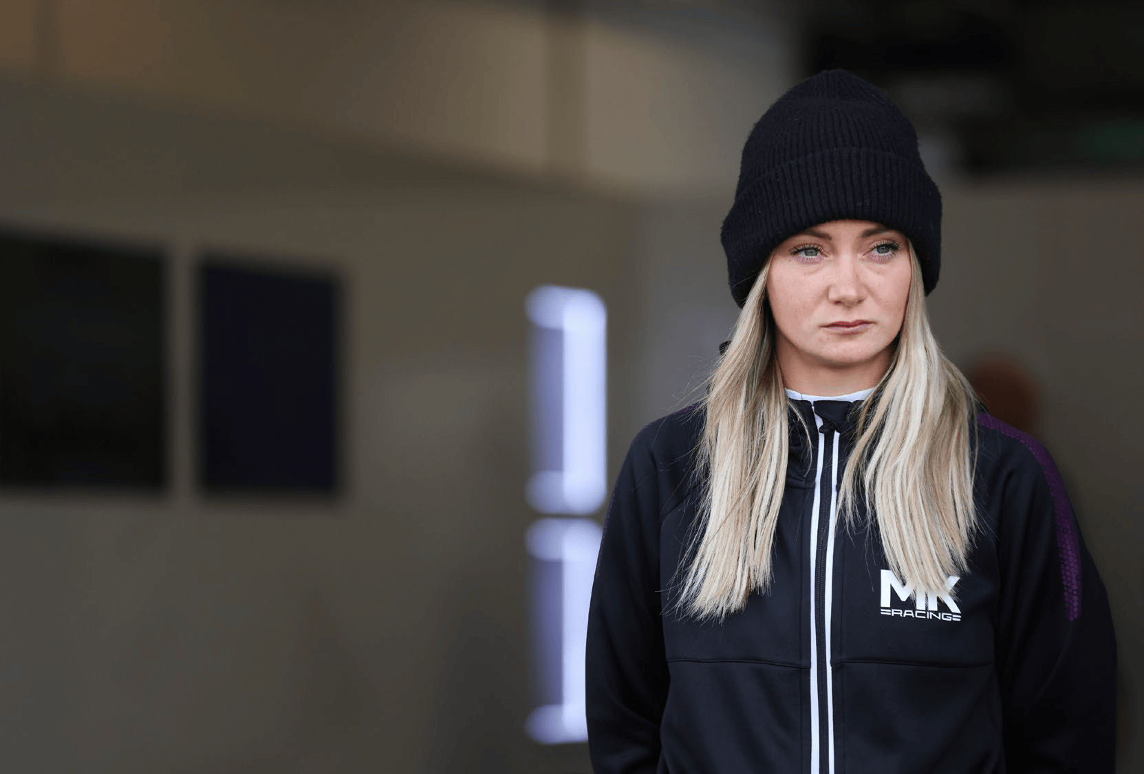 Woman with long blonde hair wearing a black beanie and black MX Racing jacket indoors.