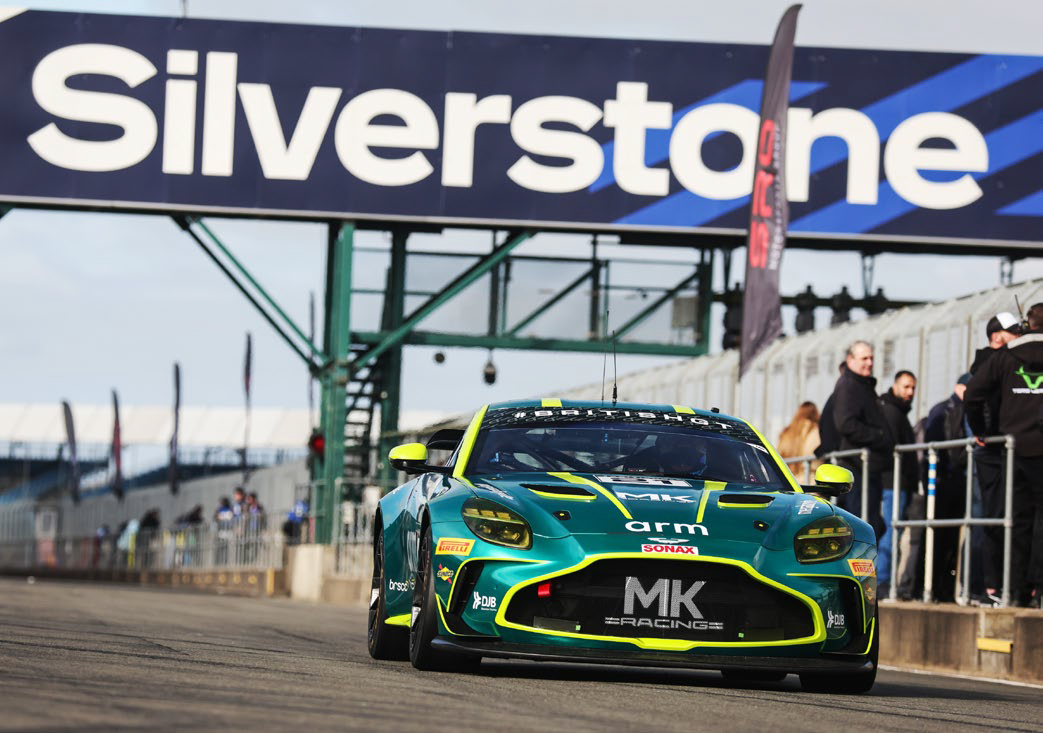 Green and yellow MK Racing car on track under Silverstone sign with people in the background.