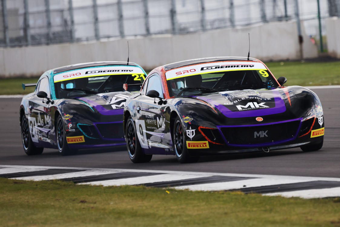 Two MK racing cars numbered 9 and 21 competing on a racetrack near a grassy curb.