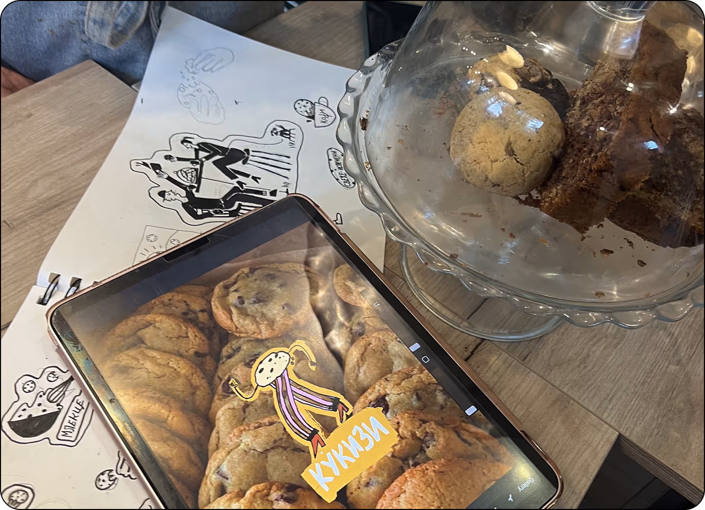 Glass cake stand holding cookies and brownies on a wooden table next to a tablet showing an image of chocolate chip cookies with a cartoon cookie character and Cyrillic text.