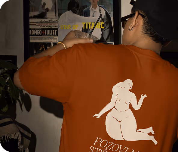 Person wearing a rusty orange t-shirt with a white abstract female figure and text on the back, holding a drink and facing a wall with framed movie posters including Romeo + Juliet and Titanic.