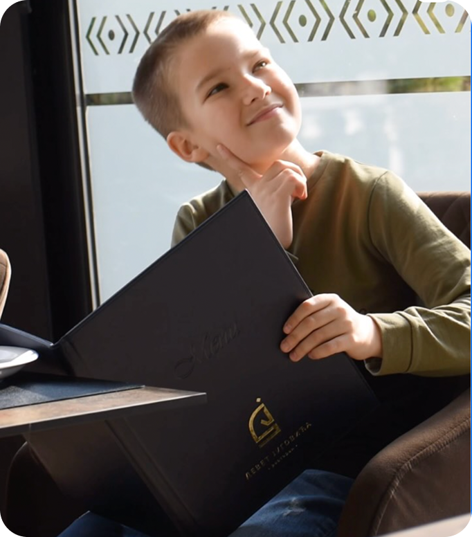 Open menu with a green page listing vitamin juices and prices in Serbian, next to a cartoon black fox wearing red boots holding a glass, on a wooden table with a person's hand nearby.