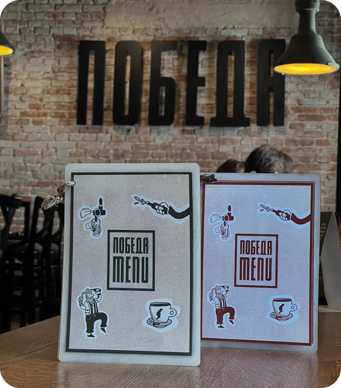 Open menu with a green page listing vitamin juices and prices in Serbian, next to a cartoon black fox wearing red boots holding a glass, on a wooden table with a person's hand nearby.