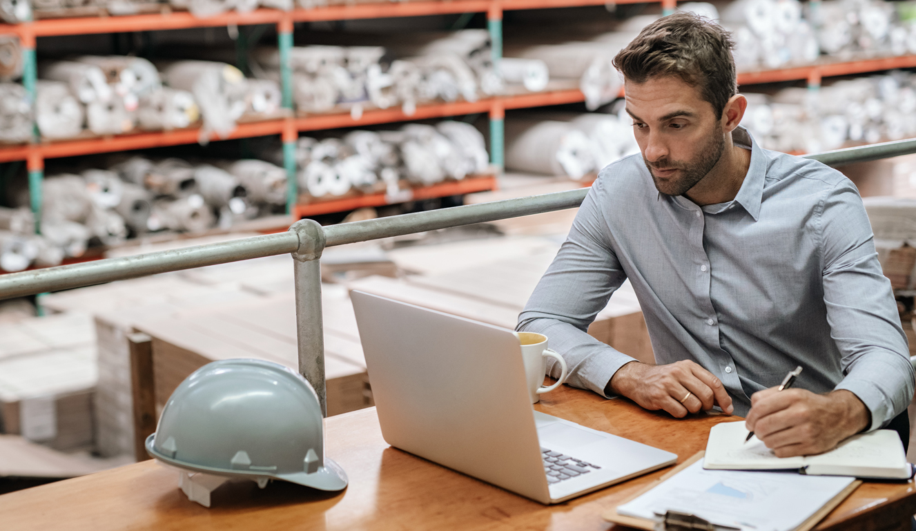 Man taking notes on notepad while looking at open laptop on table next to hard hat in warehouse