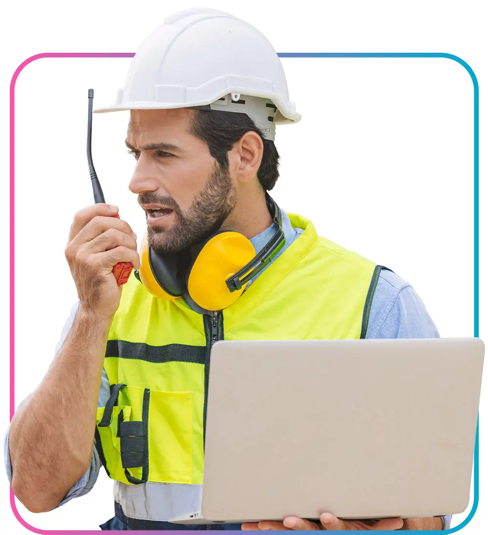 Man in hard hat and safety vest holding open laptop in one hand while talking into a 2-way radio