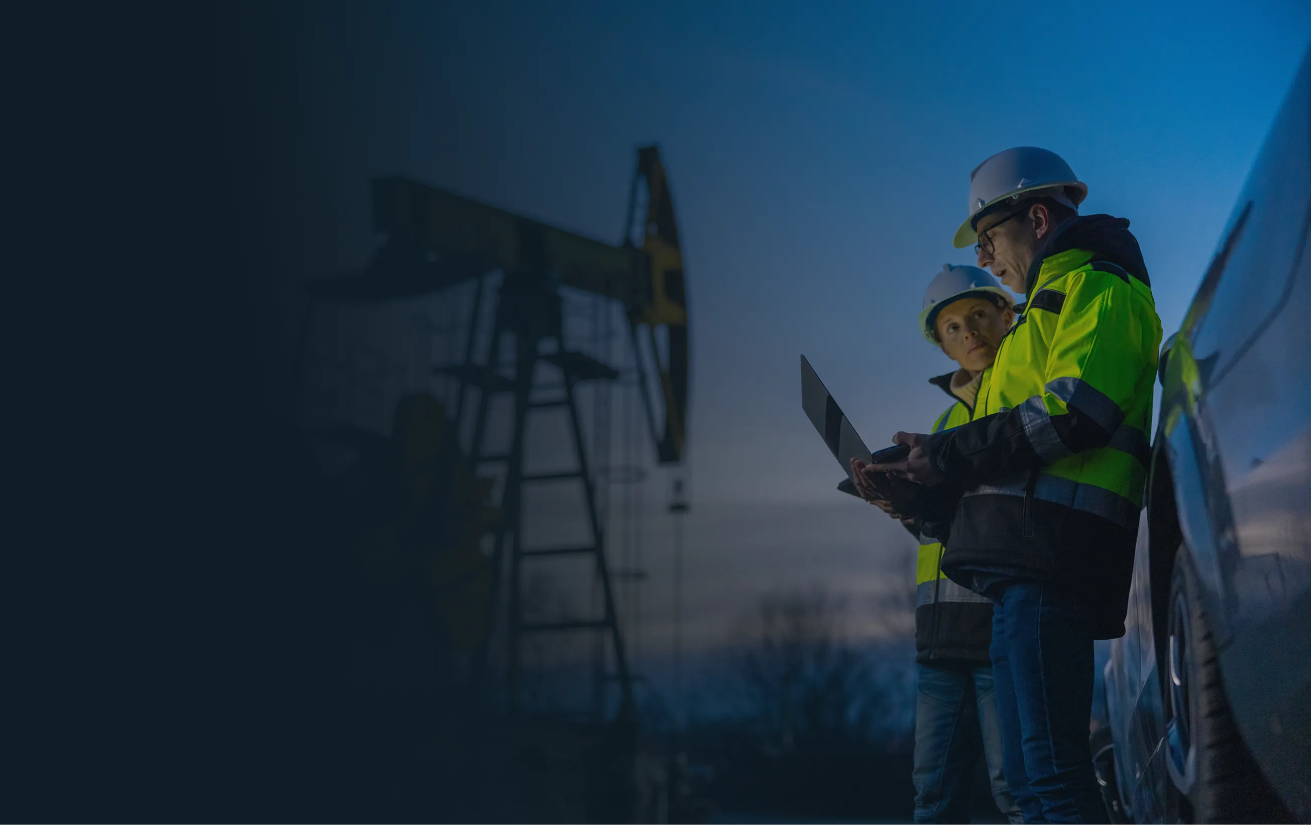 Two oilfield workers in helmets and jackets review data on a laptop near an oil pumpjack at dusk.