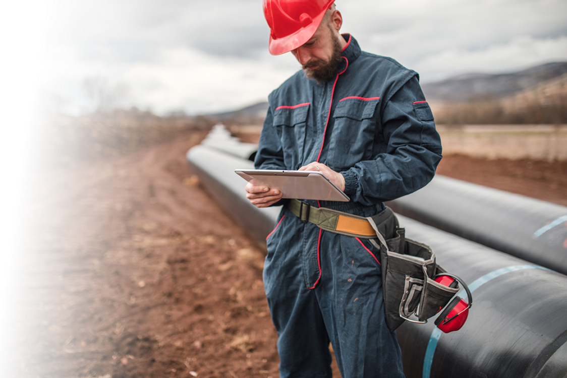 Worker in blue coveralls and red hard hat using a tablet next to large industrial pipes in a muddy outdoor setting with cloudy sky.
