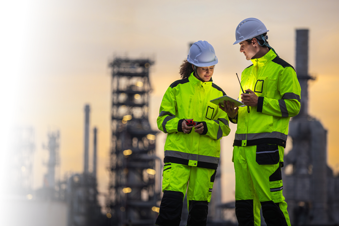 Two workers in high-visibility yellow safety gear and white helmets stand at an industrial oil refinery at sunset, one holding a tablet and the other holding a radio device.