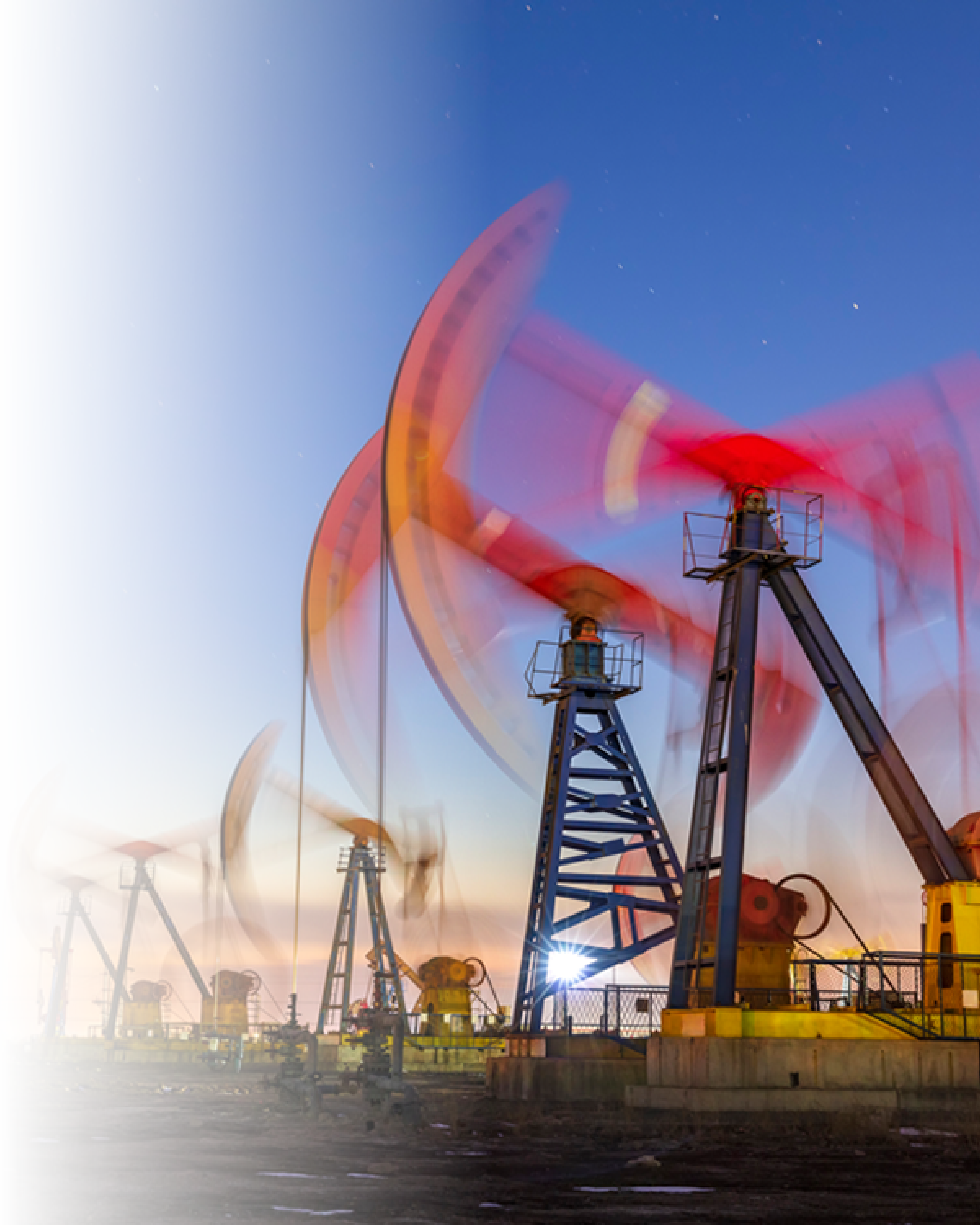 Multiple oil pumpjacks operating at dusk with motion blur on the red counterweights against a clear sky.