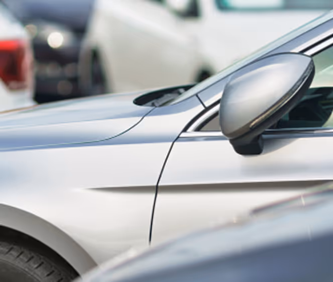 Nahaufnahme eines silbernen Autos mit Fokus auf den Seitenspiegel und die Fahrertür, im Hintergrund sind weitere geparkte Fahrzeuge unscharf zu sehen.