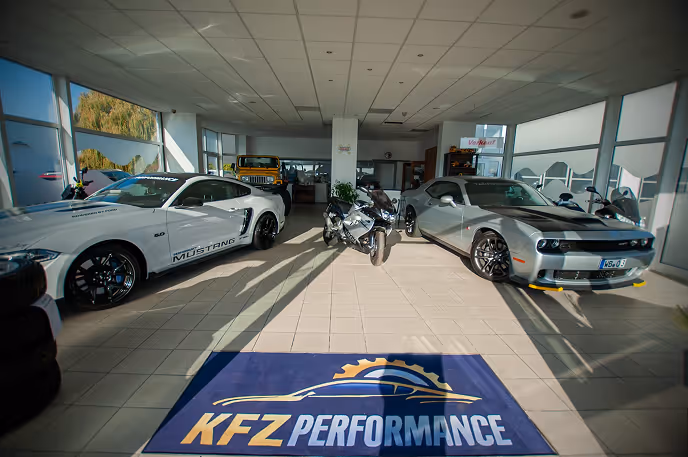 Innenraum eines Autohauses mit einem weißen Ford Mustang, einem silbernen Dodge Challenger und einem Motorrad, mit einem Teppich mit Aufschrift KFZ Performance im Vordergrund.